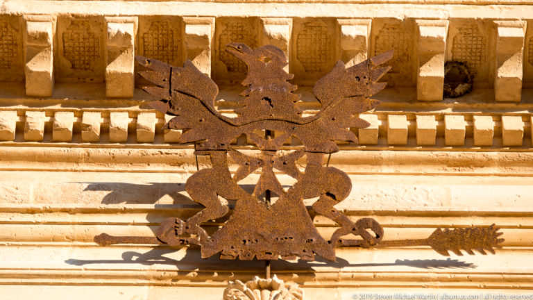 Rust-colored metal double-headed eagle emblem mounted on a pale carved stone façade beneath a projecting cornice.