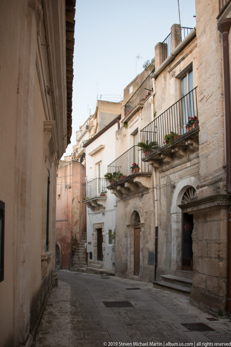 Quiet stone-paved Ragusa lane lined with pale historic houses, wrought-iron balconies, shutters, flower boxes, and carved doorways.