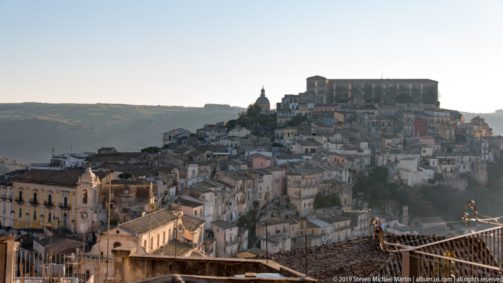 Elevated view of Ragusa Ibla with tightly packed houses, a central dome, and a large rectangular building crowning the ridge.