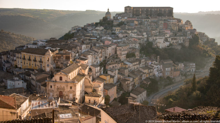 Ragusa Ibla hillside with a church in the foreground and densely layered buildings climbing toward the ridge.