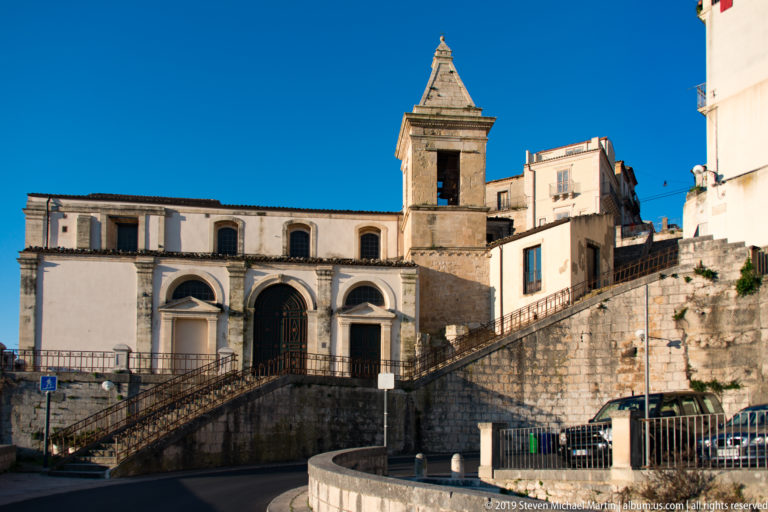 Low-angle view of a pale stone church and square bell tower above a broad exterior stair and retaining wall.