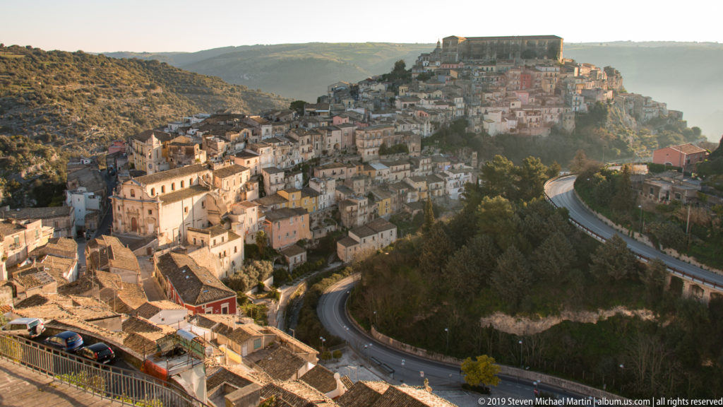 Panoramic view of Ragusa Ibla climbing a hill above a curving road and wooded ravine in warm late-day light.