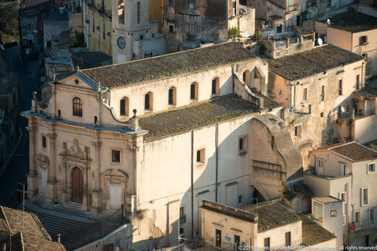 Elevated view of a large pale stone church with tiled roof, arched windows, bell tower, and surrounding historic buildings.