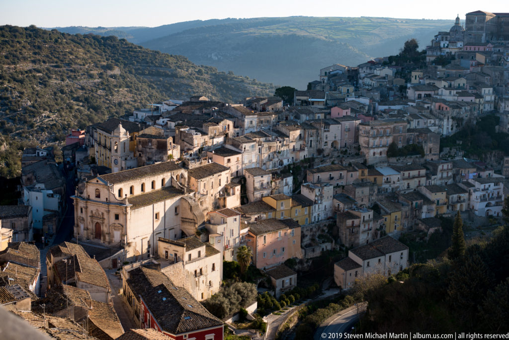 Wide hillside view of Ragusa with tightly packed pale buildings, a church in the foreground, and higher structures on the ridge.