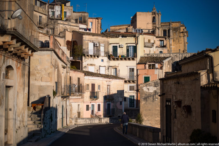 Narrow historic street lined with weathered buildings and balconies, leading uphill toward a church under a deep blue sky.