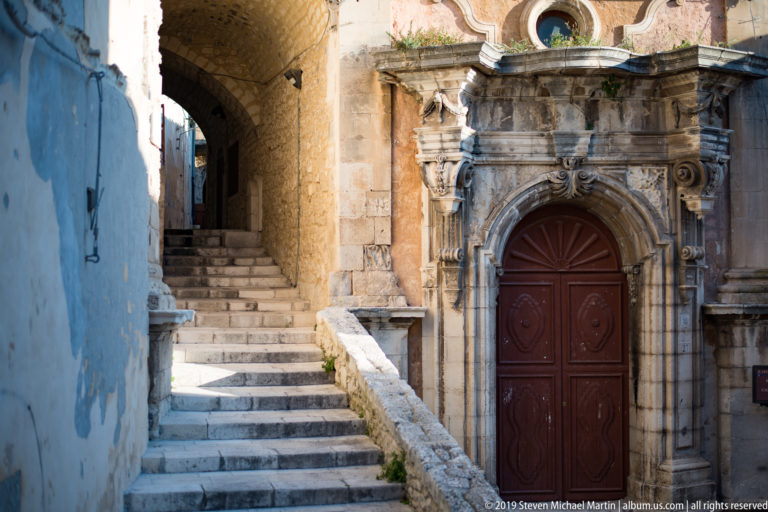 Ornate stone church portal with a red double door beside a narrow flight of steps passing under a masonry arch.