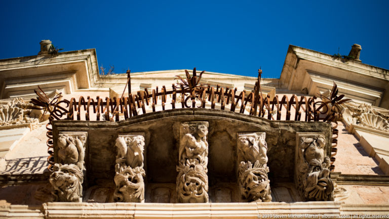 Low-angle view of a stone balcony supported by carved human and grotesque corbels beneath iron railings.
