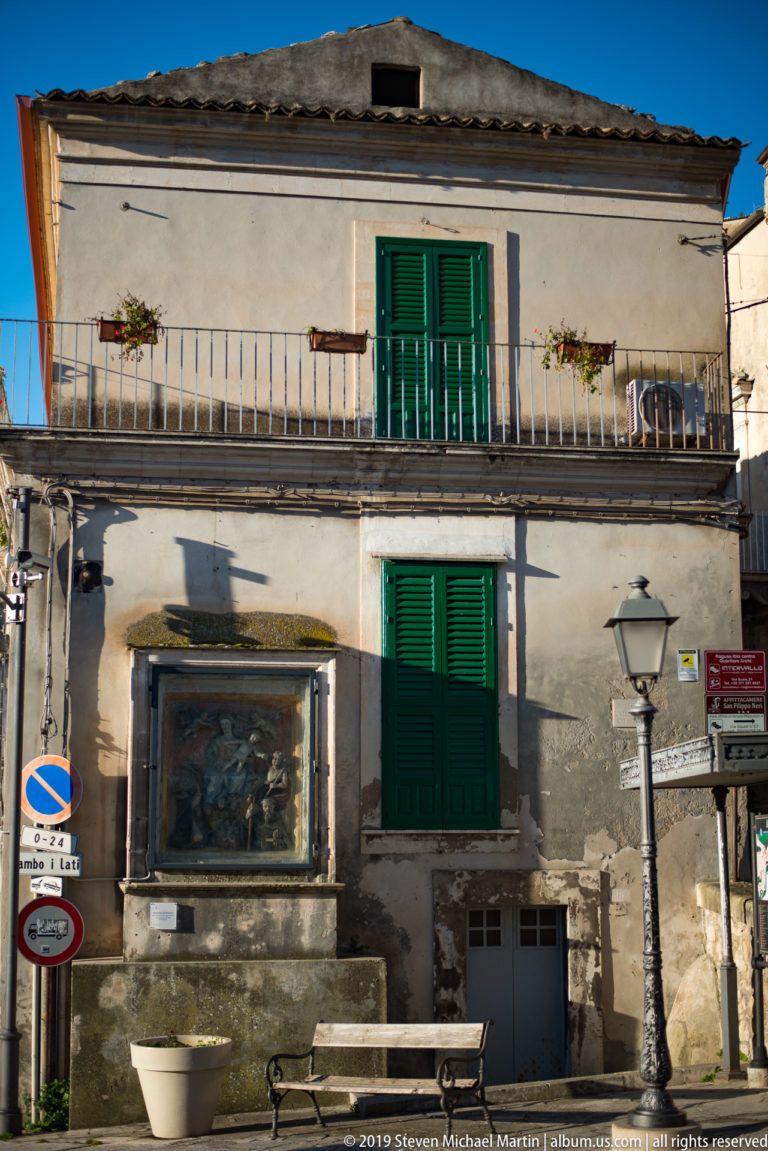 Weathered house with green shutters, long balcony, framed wall panel, benches, and street lamp.