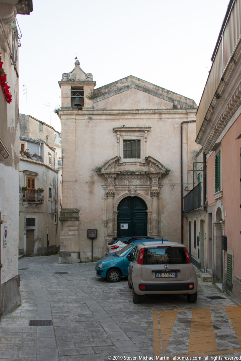Small gabled church with ornate central stone portal and dark arched door facing a paved square with parked cars.