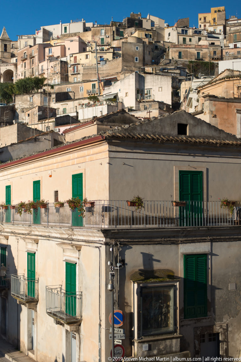 Sunlit house with green shutters and balcony before densely layered buildings climbing a Ragusa hillside.