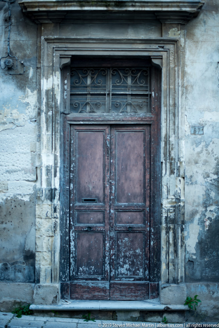 Weathered dark wooden double door with paneled leaves, decorative transom grille, and a projecting carved stone surround.