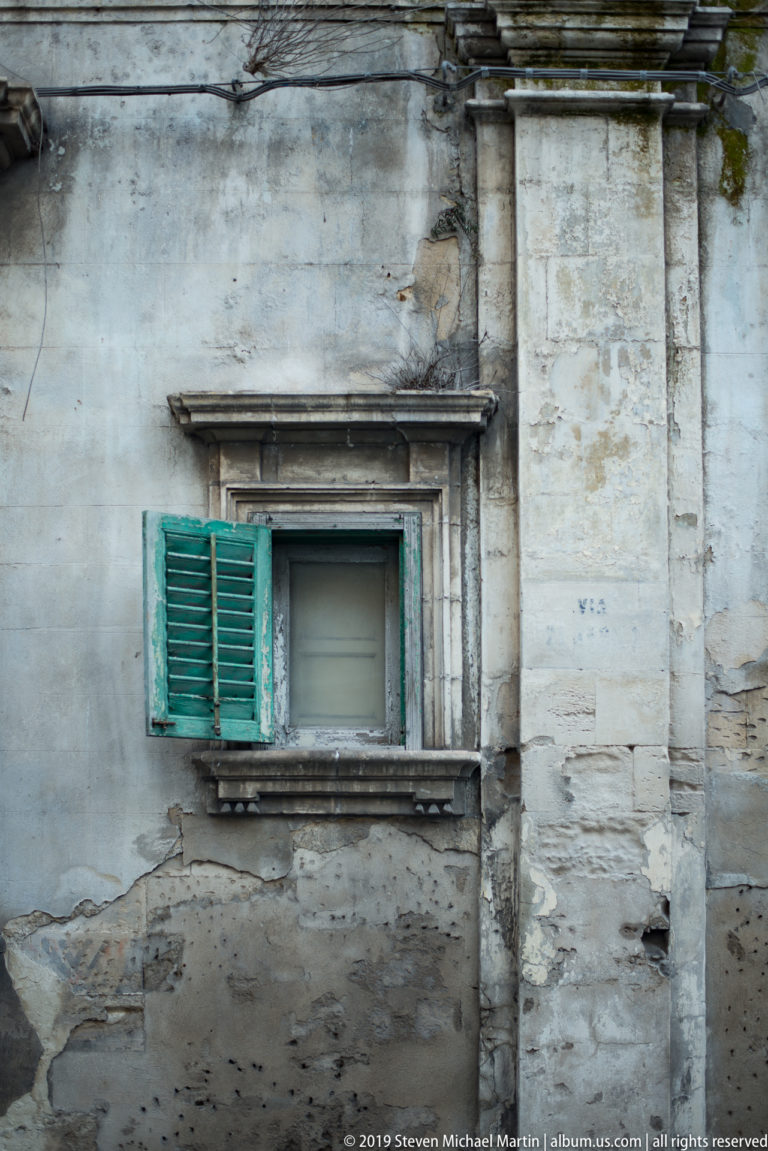 Small green-shuttered window set in a stained, weathered stone wall beside a heavy vertical pier.