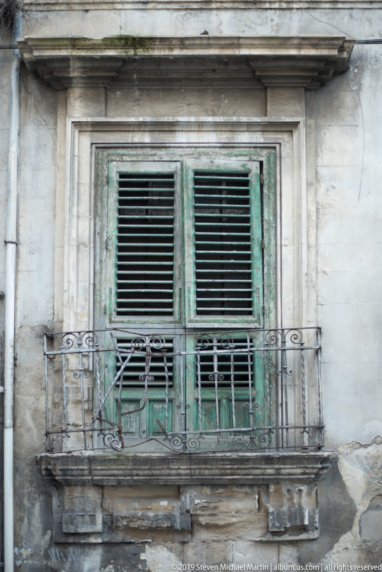 Weathered green louvered shutters behind a wrought-iron balcony on a cracked pale stone façade.