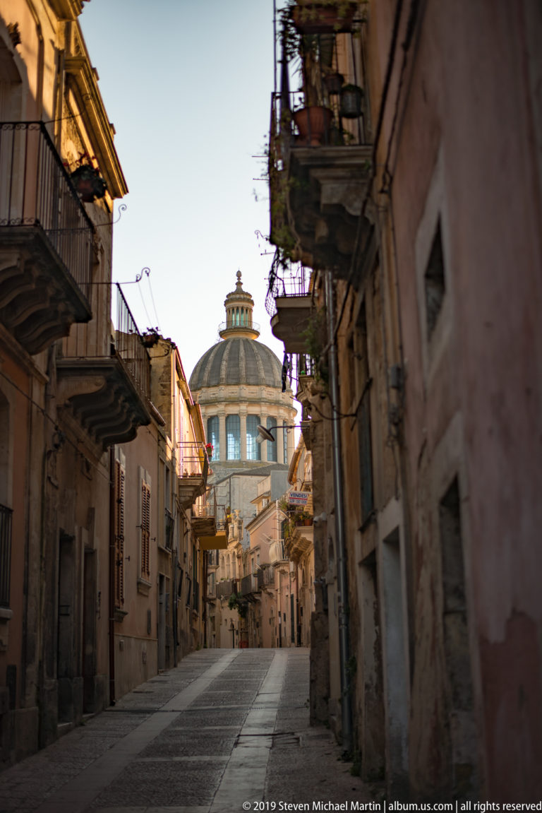 Quiet stone-paved Ragusa Ibla street with balconies and the dome of San Giorgio rising in the distance.
