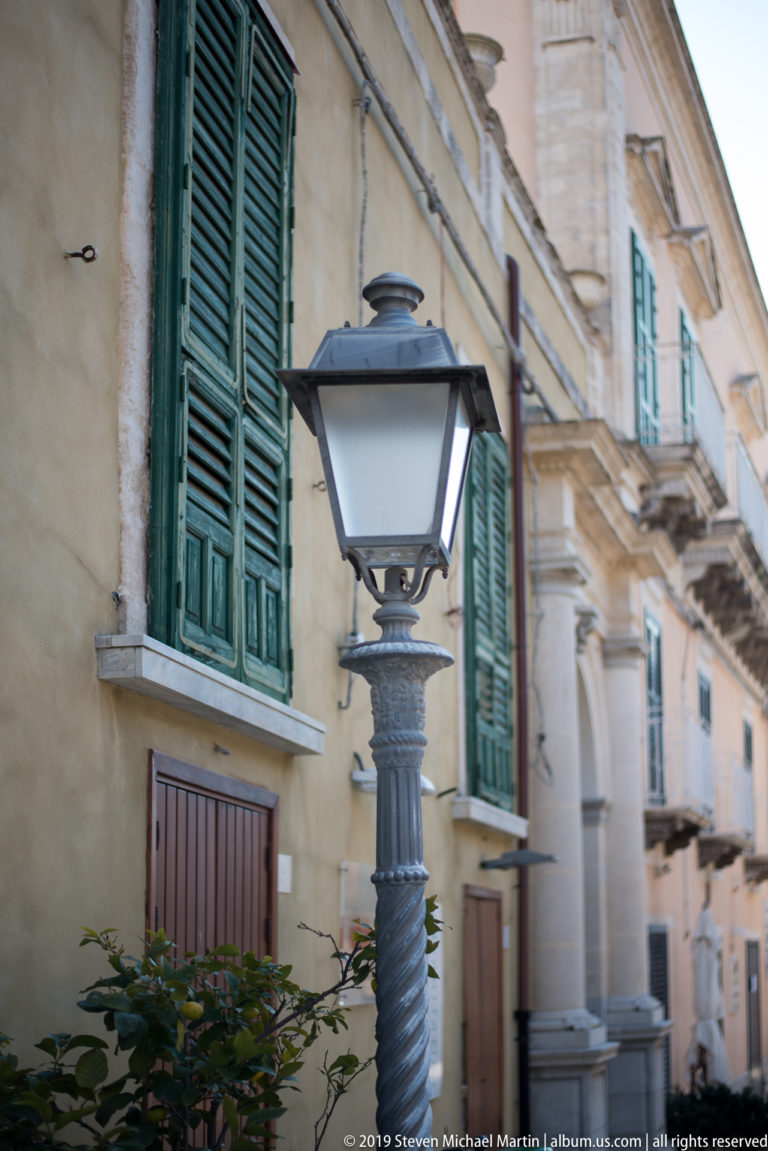 Black decorative street lamp before a weathered building with green shutters and balconies in Ragusa.