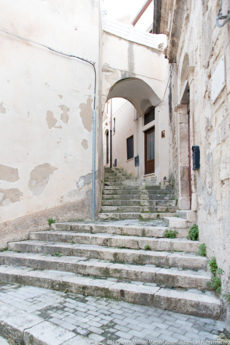 Worn stone steps rising beneath a shallow connecting arch between weathered plaster and masonry buildings.
