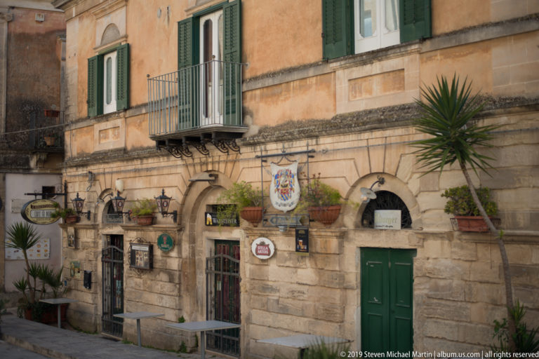 Weathered stone building with arched storefronts, green shutters, balconies, flower pots, signs, and a crest.