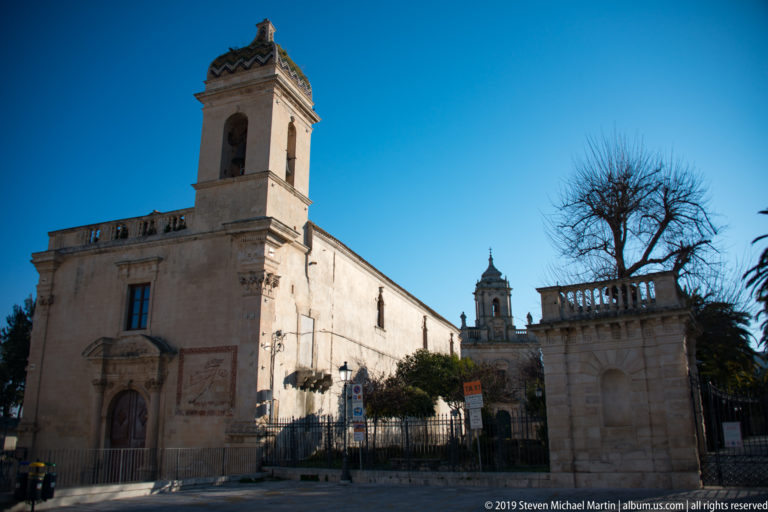 Pale stone church with square bell tower, side-wall sundial, and balustraded terrace under blue sky.