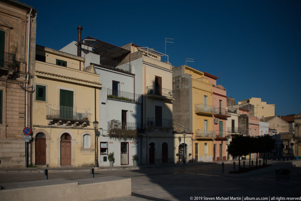 Quiet Ragusa square or street edge lined with pastel houses, balconies, shutters, and rooftop terraces.