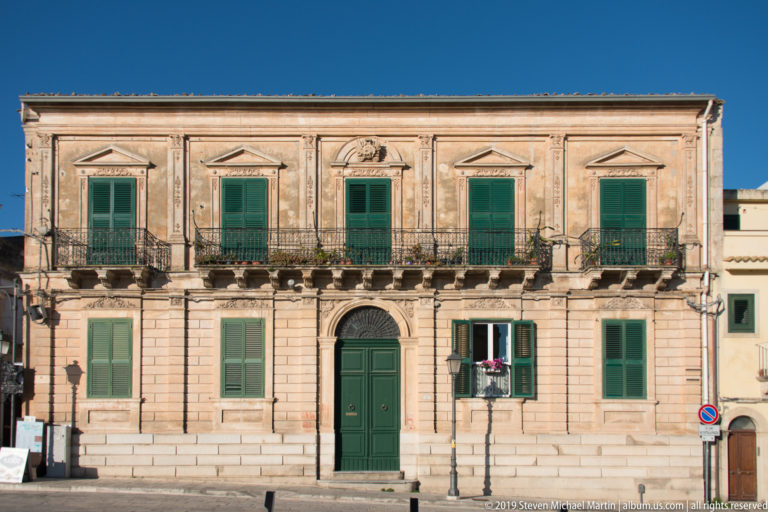 Two-story stone palazzo with central arched green door, green shutters, pediments, and iron balconies.
