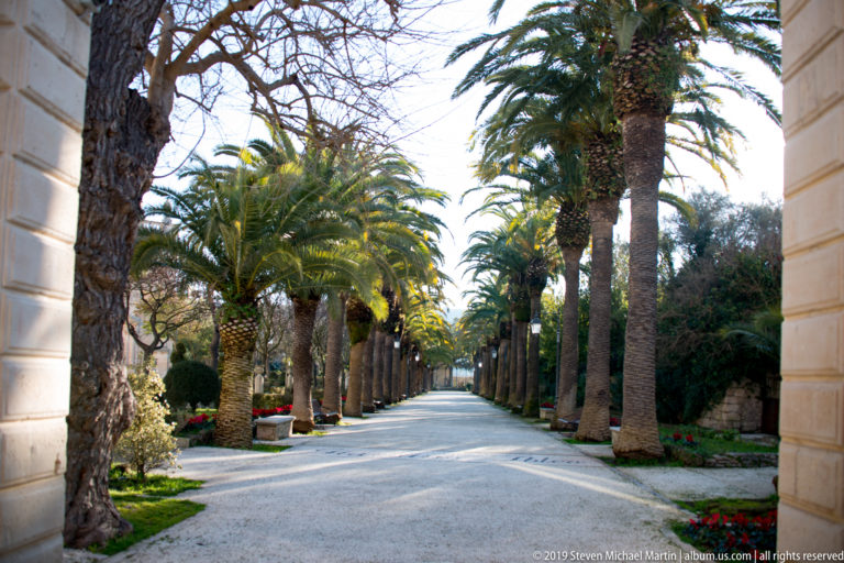 Straight garden promenade lined with tall palms, clipped shrubs, benches, and flower beds in Ragusa.