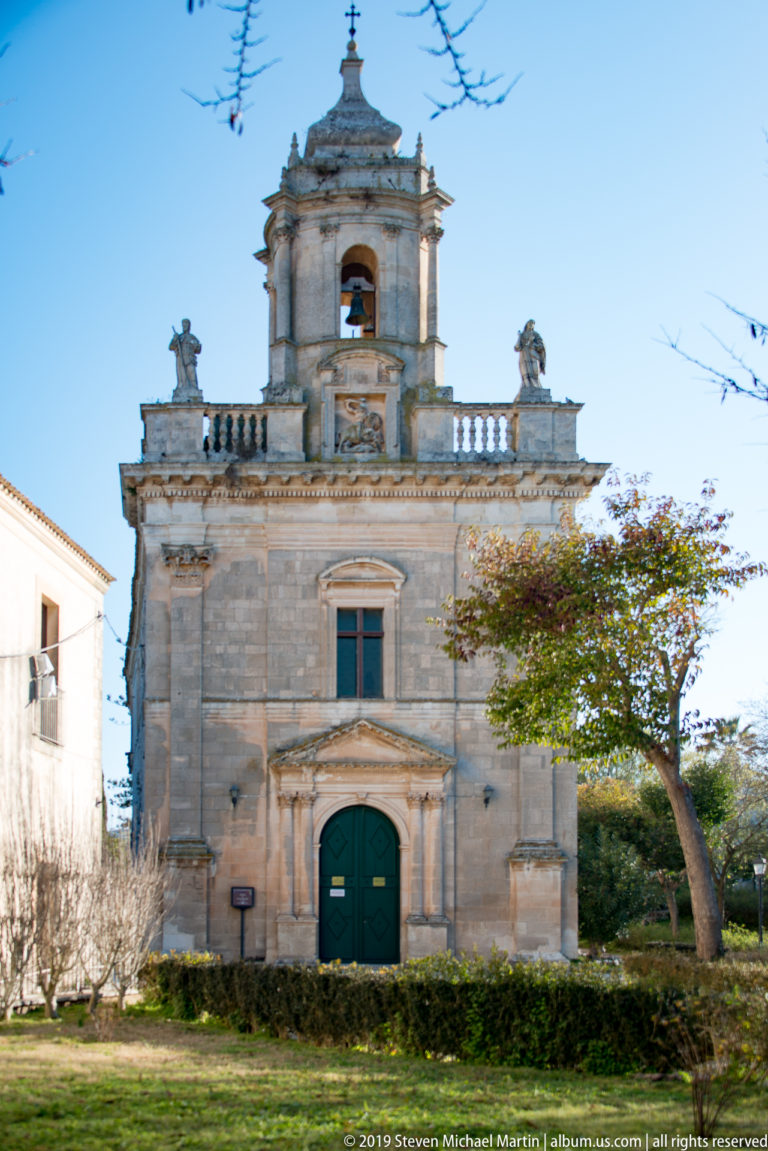 Small pale-stone Baroque church with green door, upper bell opening, balustrade, and cross.