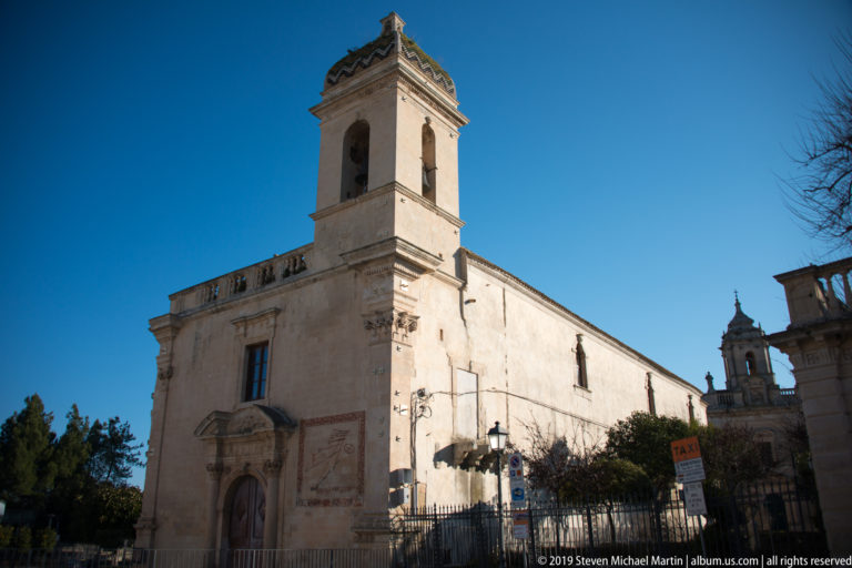 Sunlit pale stone church with square bell tower, side-wall sundial, and garden wall beneath blue sky.