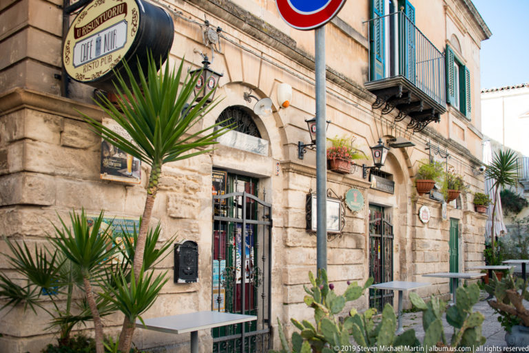 Rusticated stone storefront with green shutters, iron balconies, potted palms, cactus, and outdoor tables.