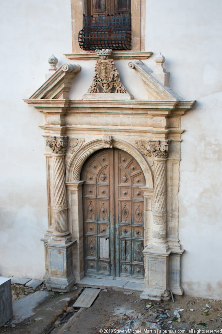 Weathered wooden double door framed by an ornate stone portal with twisted columns, carved capitals, broken pediment, crest, and balcony above.