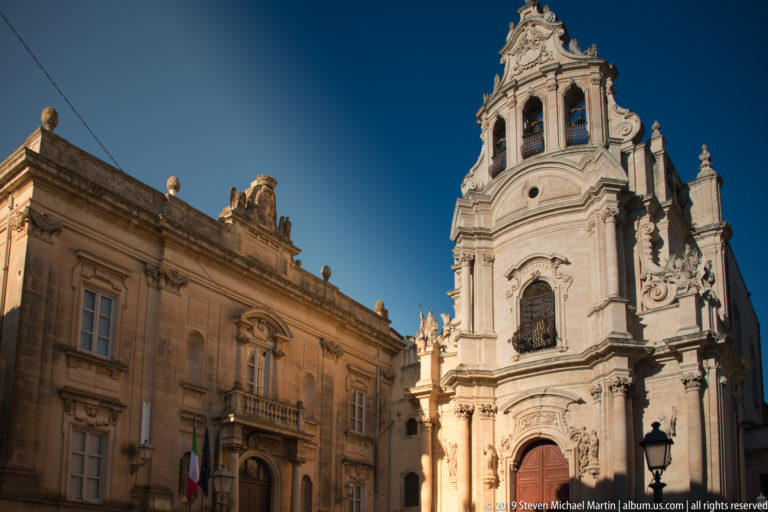 Ornate pale-stone Baroque church with bell stage beside a formal historic palazzo under blue sky.