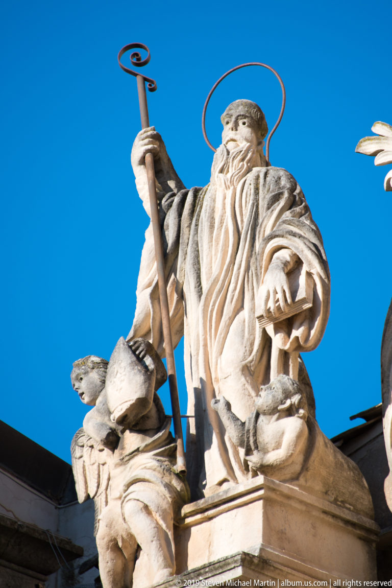 Low-angle view of a bearded bishop saint holding a crozier, with a metal halo against blue sky.