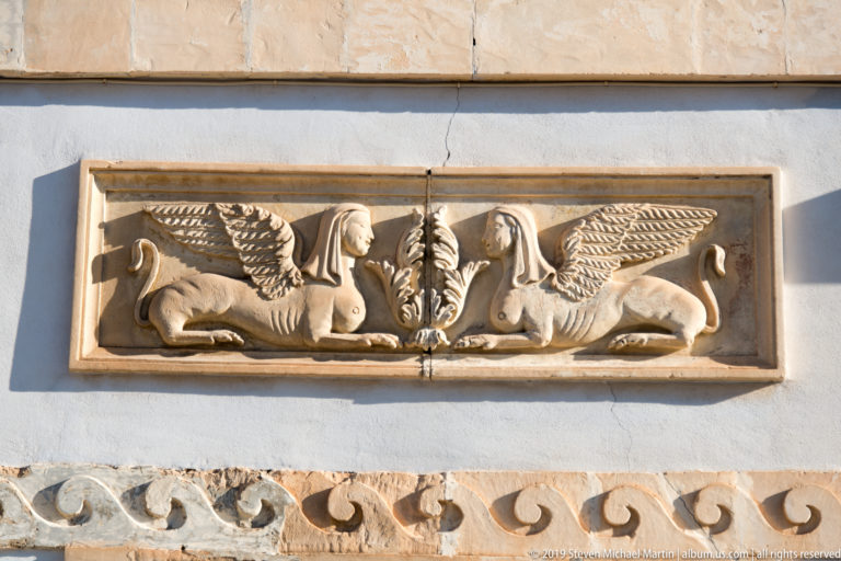 Stone wall relief of two winged leonine creatures facing a central plant motif above a carved scroll band.