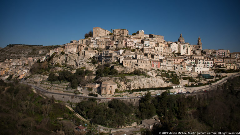 Ragusa Ibla descending a steep hillside above a curving road and wooded ravine in bright sunlight.