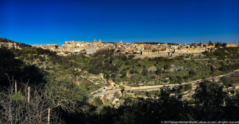 Sunlit Ragusa buildings and church towers spread across a ridge beyond a deep green valley.