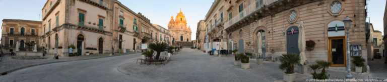 Quiet stone street in Ragusa Ibla lined with historic buildings and palms, with a sunlit bell tower ahead.