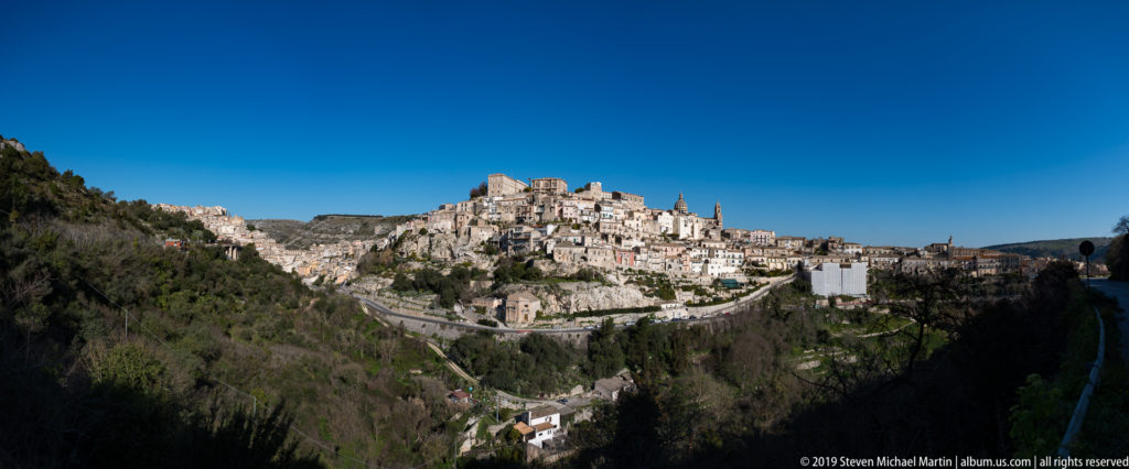 Ragusa Ibla clustered on a steep ridge above a green ravine under a deep blue sky.