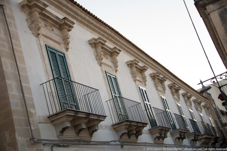 Oblique view along a pale historic façade with repeated green-shuttered windows, wrought-iron balconies, and carved stone supports.