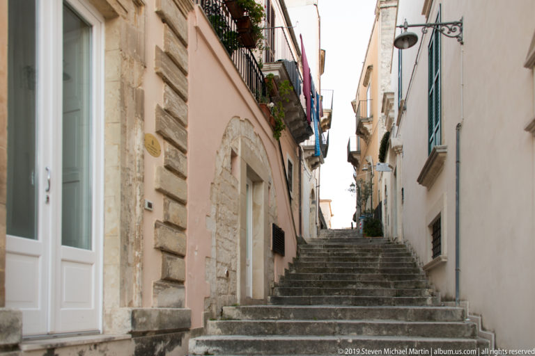 Steep worn stone stairway rising between narrow residential buildings with balconies, exposed masonry, utility lines, and hanging laundry.