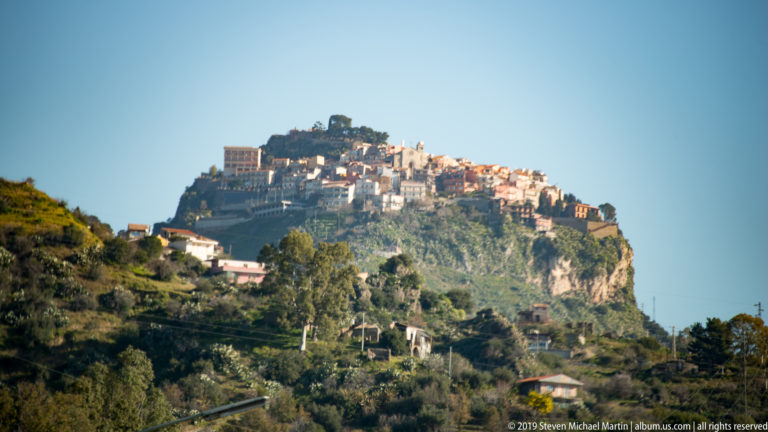 Warm-toned hill town clustered atop a steep rocky summit, with wooded slopes and exposed cliffs below.