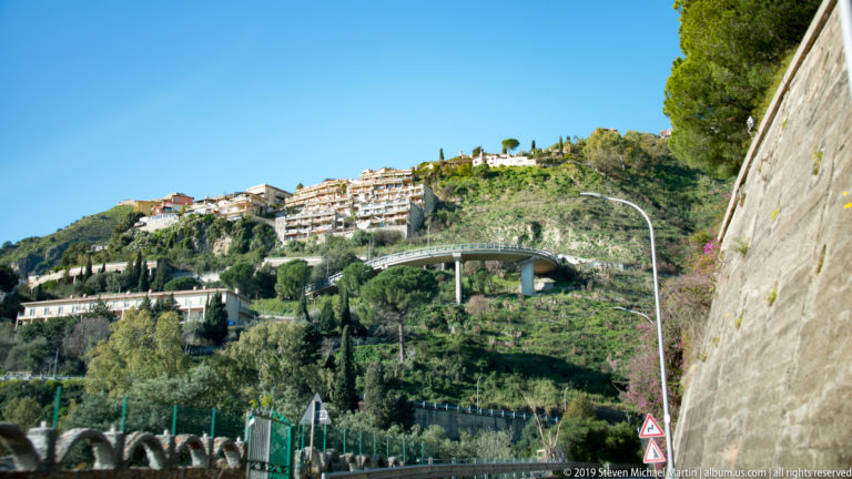 Road beside a tall retaining wall facing a curving elevated roadway and apartment buildings on a green hillside.