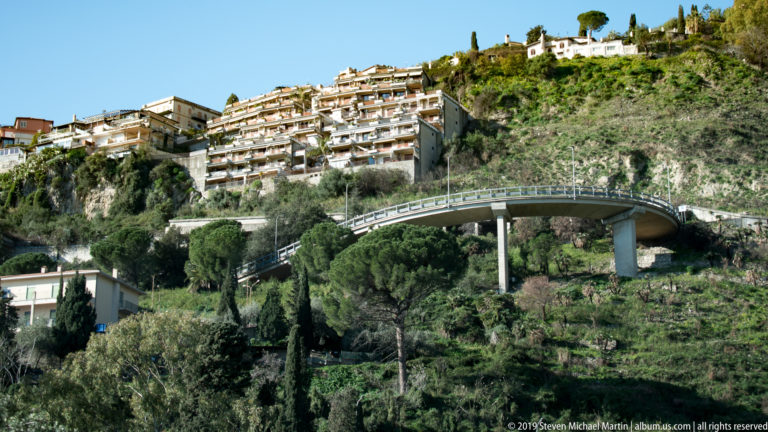 Curving elevated road on concrete piers below apartment buildings and terraced green hillside vegetation.