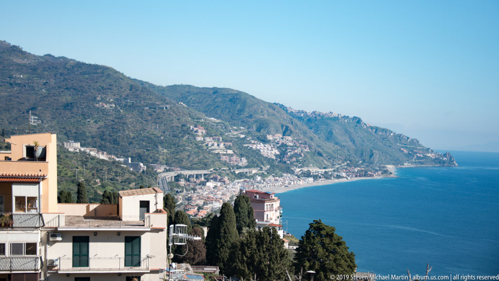 Taormina hillside buildings overlooking a long mountainous coastline lined with towns beside the blue Ionian Sea.