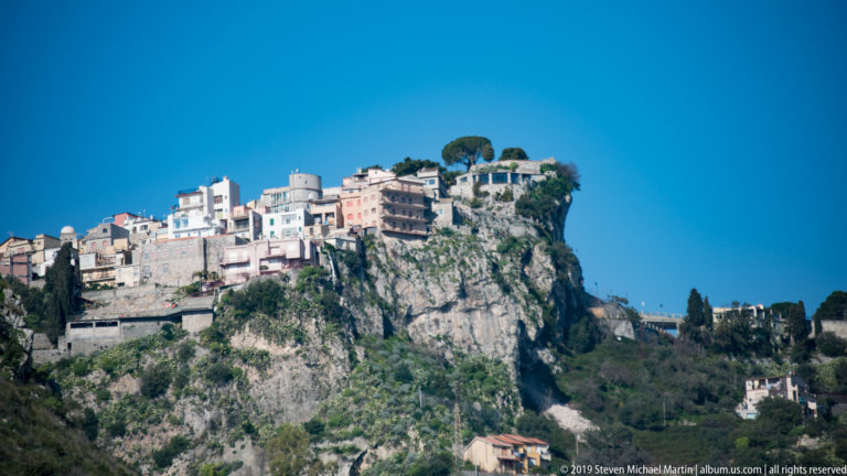 Compact hilltop town clustered on a steep rocky summit above green slopes under a clear blue sky.