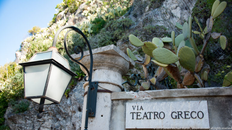 Stone Via Teatro Greco street sign beneath a black lantern, with prickly pear cactus growing against rock behind it.