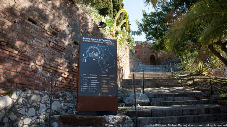 Stone steps climb beside a theatre orientation sign, brick wall, palms, and other Mediterranean plants.