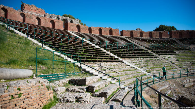 Curving tiers of stone and wooden theatre seating with stairs, railings, and a visitor near the lower aisle.