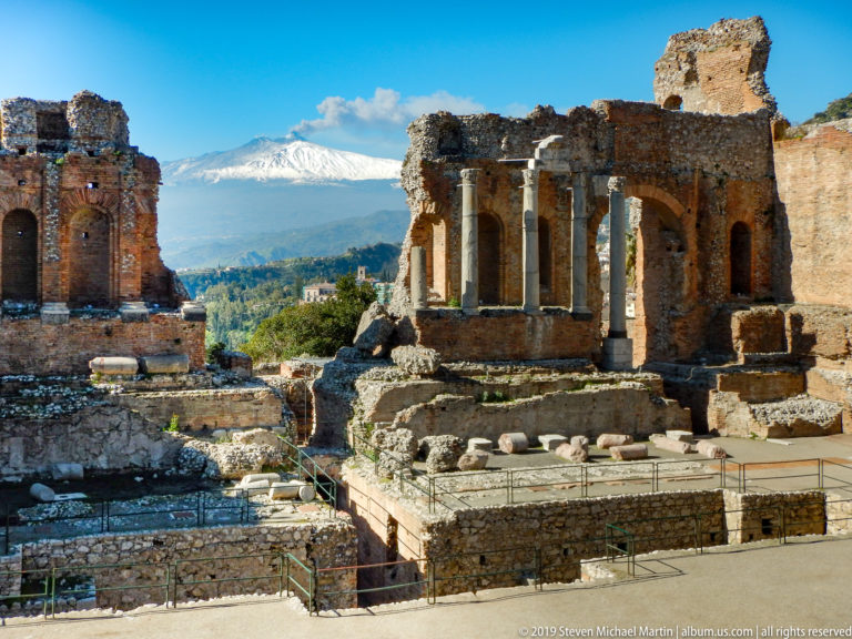 Standing columns, brick stage ruins, and excavated foundations with snow-covered Mount Etna visible beyond.