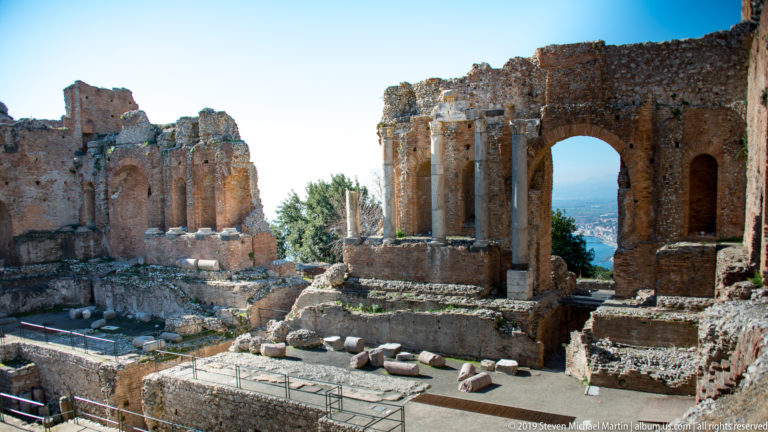 Ruined brick stage walls with columns and arches separated by an open gap above stone foundations.
