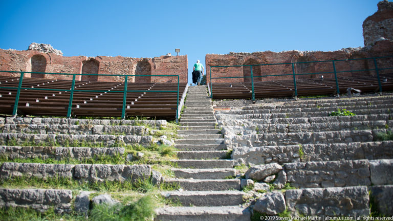 Tiered stone theatre seating divided by a central stair, with one visitor climbing toward the ruined upper wall.