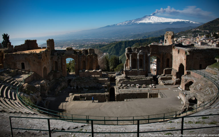Brick stage ruins and columns of the ancient theatre with snow-covered Mount Etna rising behind them.
